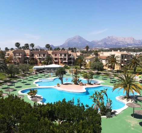 Resort con piscina rodeada de palmeras y monta&ntilde;as al fondo bajo cielo azul.