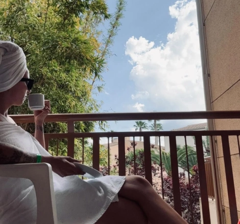 Persona sentada en balc&oacute;n, tomando caf&eacute;, con toalla en la cabeza y cielo despejado.