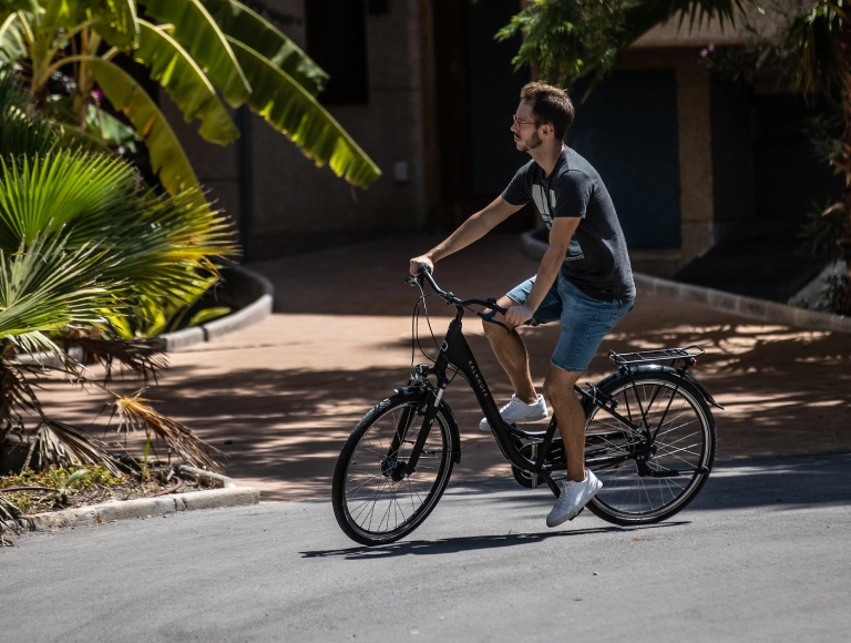 Persona montando bicicleta en entorno urbano, rodeado de plantas tropicales.