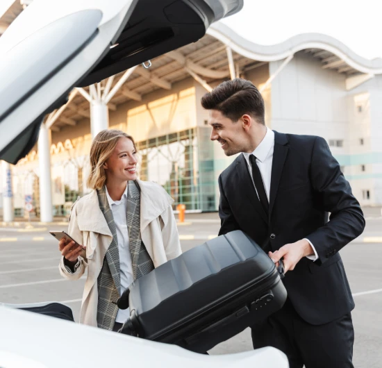 Hombre y mujer sonr&iacute;en junto a auto con maleta en aeropuerto.