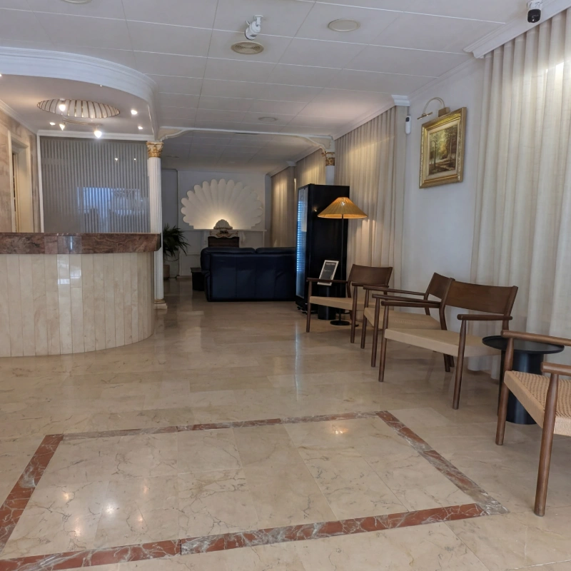 Recepci&oacute;n de hotel con mostrador, sof&aacute; y zona de espera con mesas y sillas.