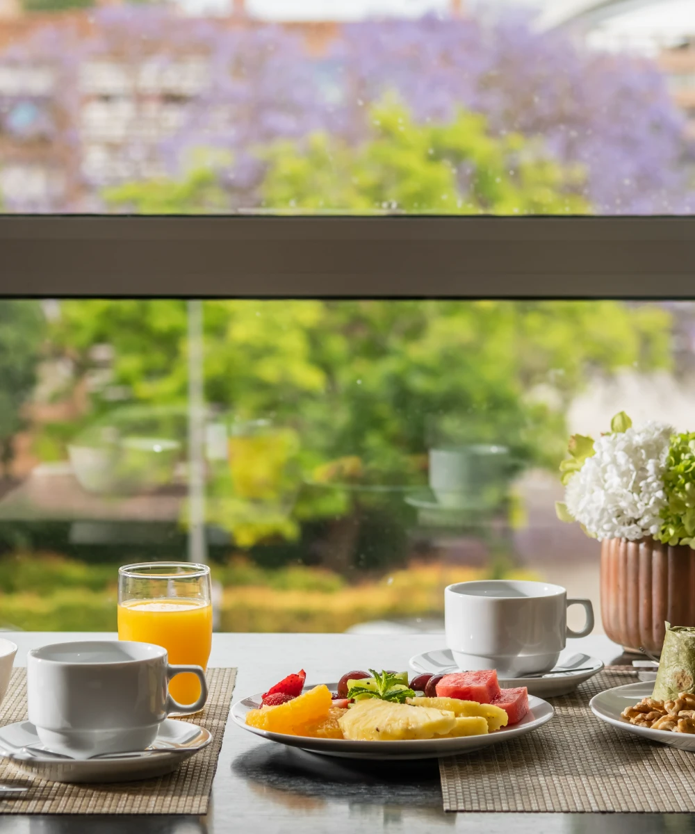 Desayuno con frutas, caf&eacute; y jugo frente a una ventana con vista a un jard&iacute;n.