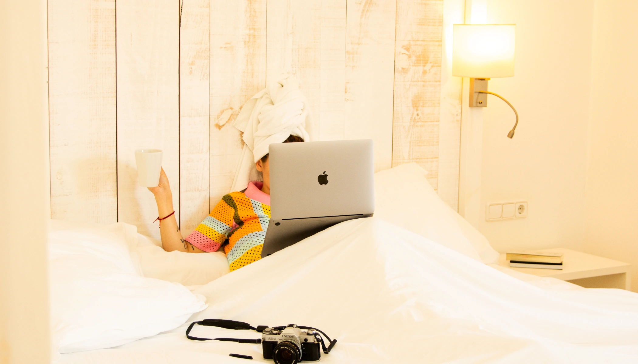 Persona en cama con laptop, taza en mano y c&aacute;mara sobre la s&aacute;bana.