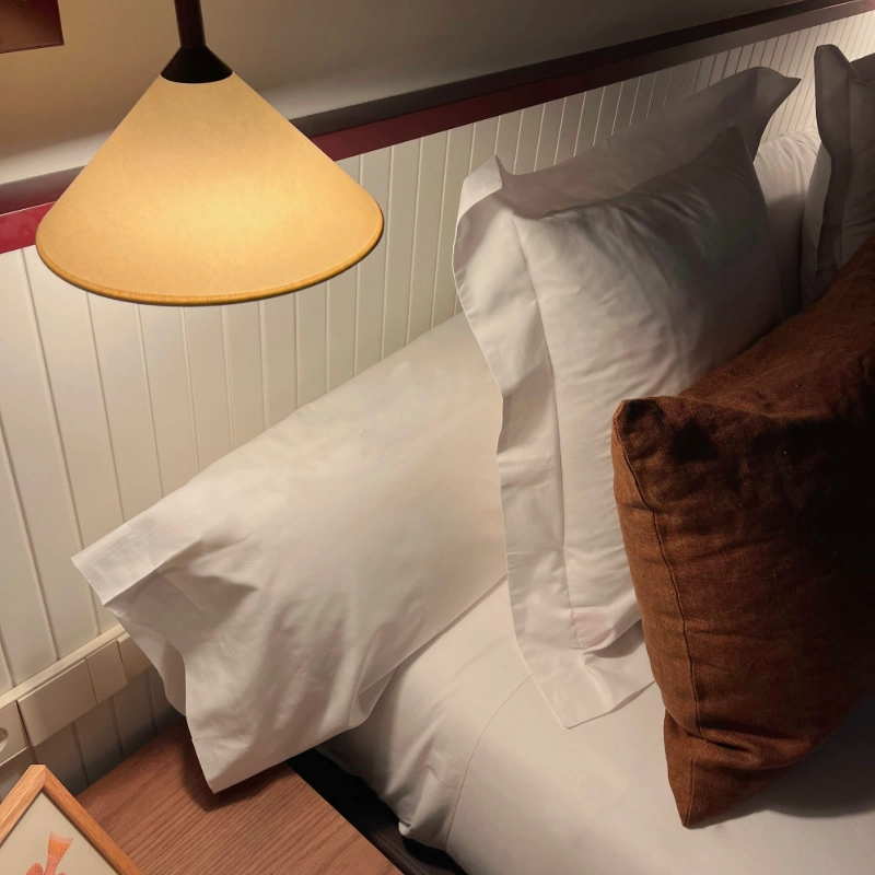 L&aacute;mpara iluminando una cama con almohadas blancas y marrones.