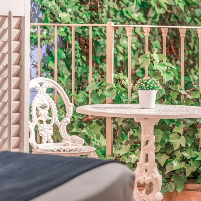 Silla y mesa de hierro blanco con planta, frente a cerca y hojas verdes.