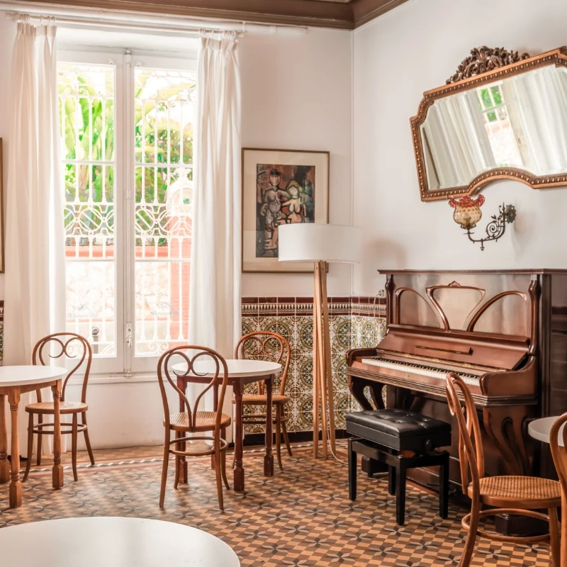 Sala con piano, mesas y sillas de madera, espejo y ventana con cortinas blancas.