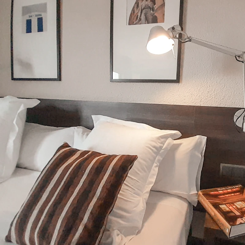 Cama con almohadas y cojín a rayas junto a una lámpara de lectura encendida.