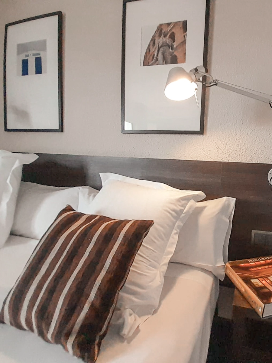 Cama con almohada rayada, l&aacute;mpara y cuadros de fondo encima del cabecero oscuro.