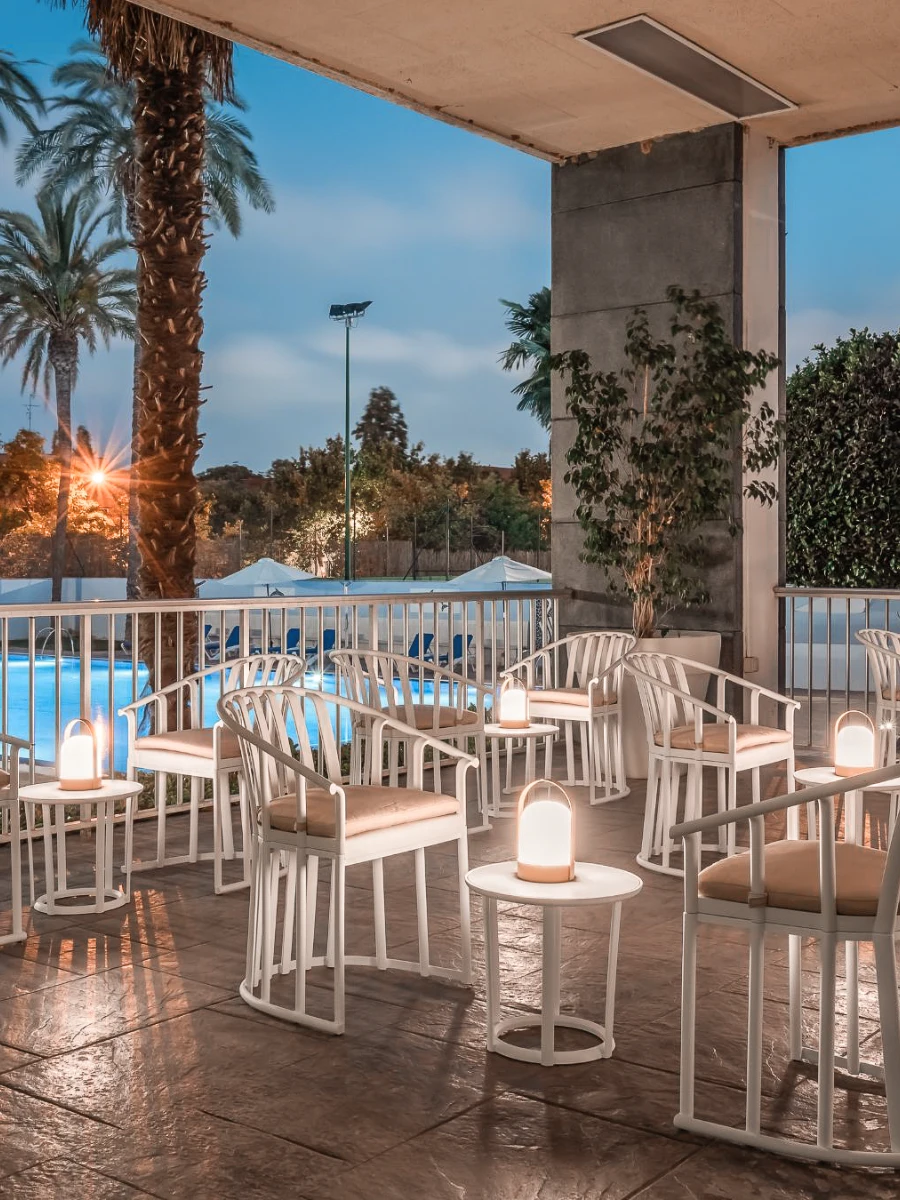Terraza con sillas blancas y mesas con l&aacute;mparas, junto a una piscina al atardecer.