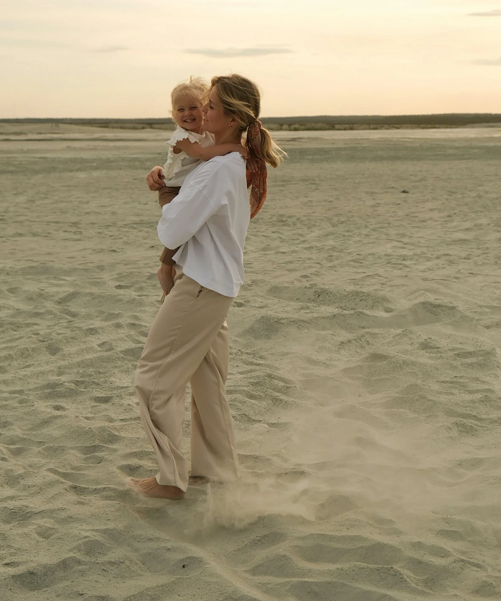 Mujer sonriente con ni&ntilde;o en brazos en una playa al atardecer.