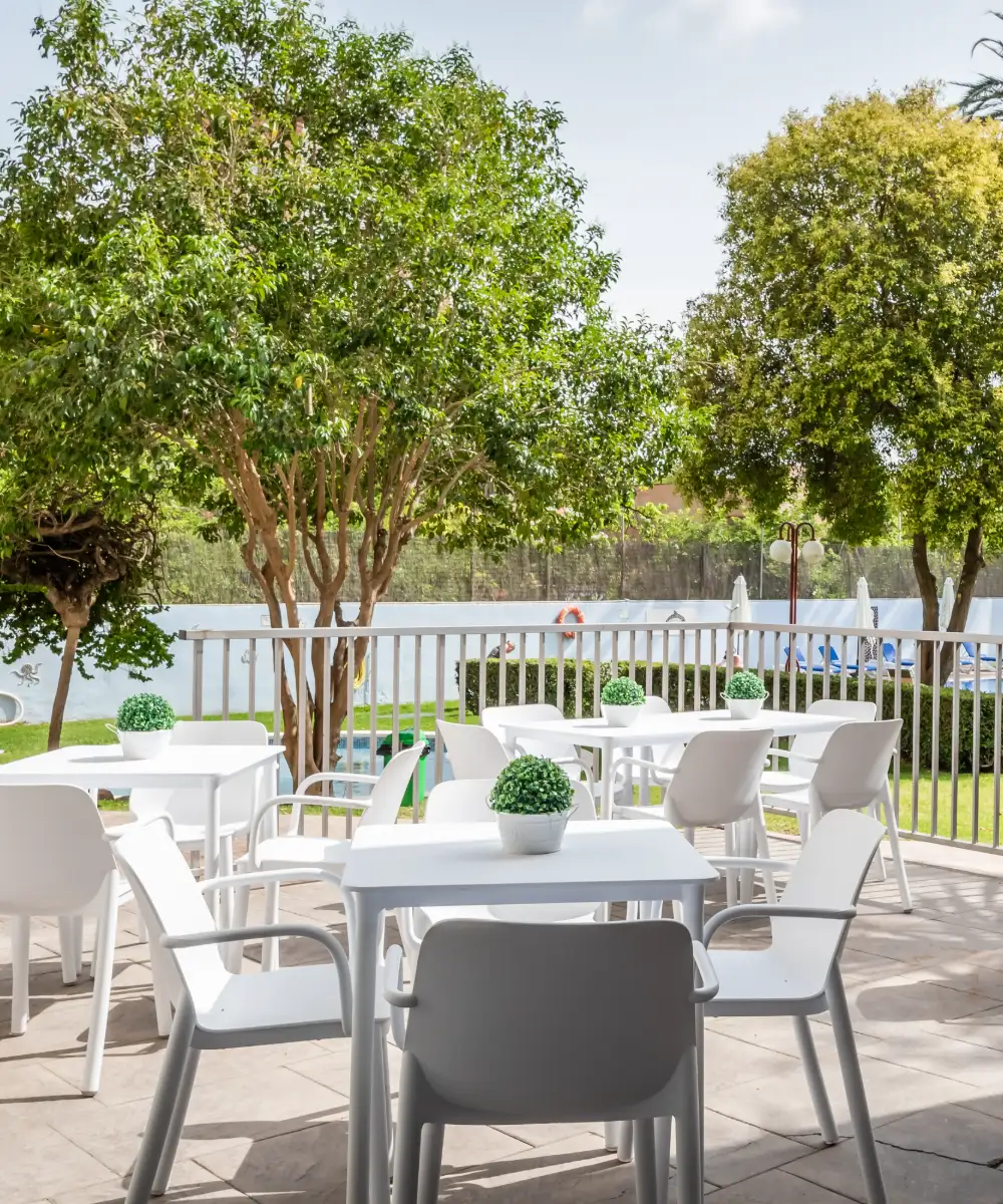 Mesas blancas en una terraza con plantas y vista al jard&iacute;n.