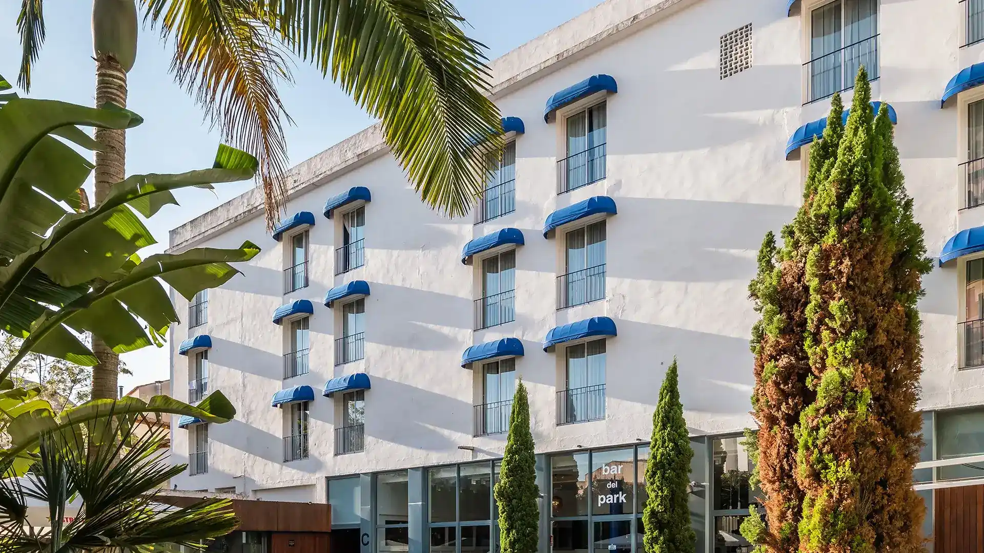Edificio blanco con ventanas, toldos azules y palmeras alrededor.