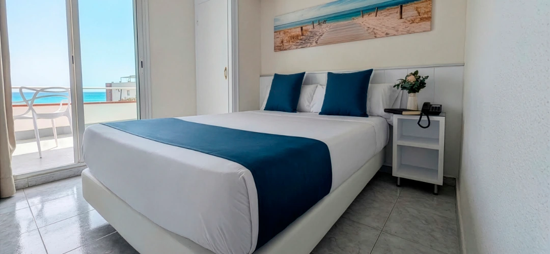 Habitaci&oacute;n luminosa con cama blanca y vista al mar desde el balc&oacute;n.