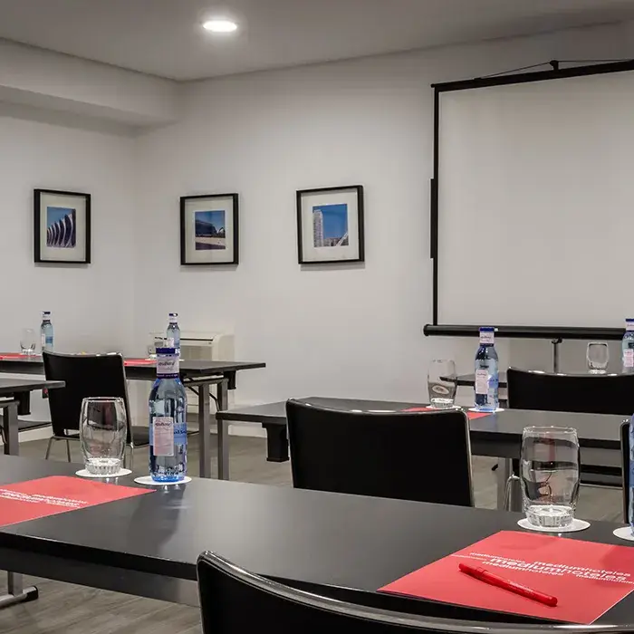 Sala de reuniones con mesas, sillas, botellas de agua y proyector en la pared.