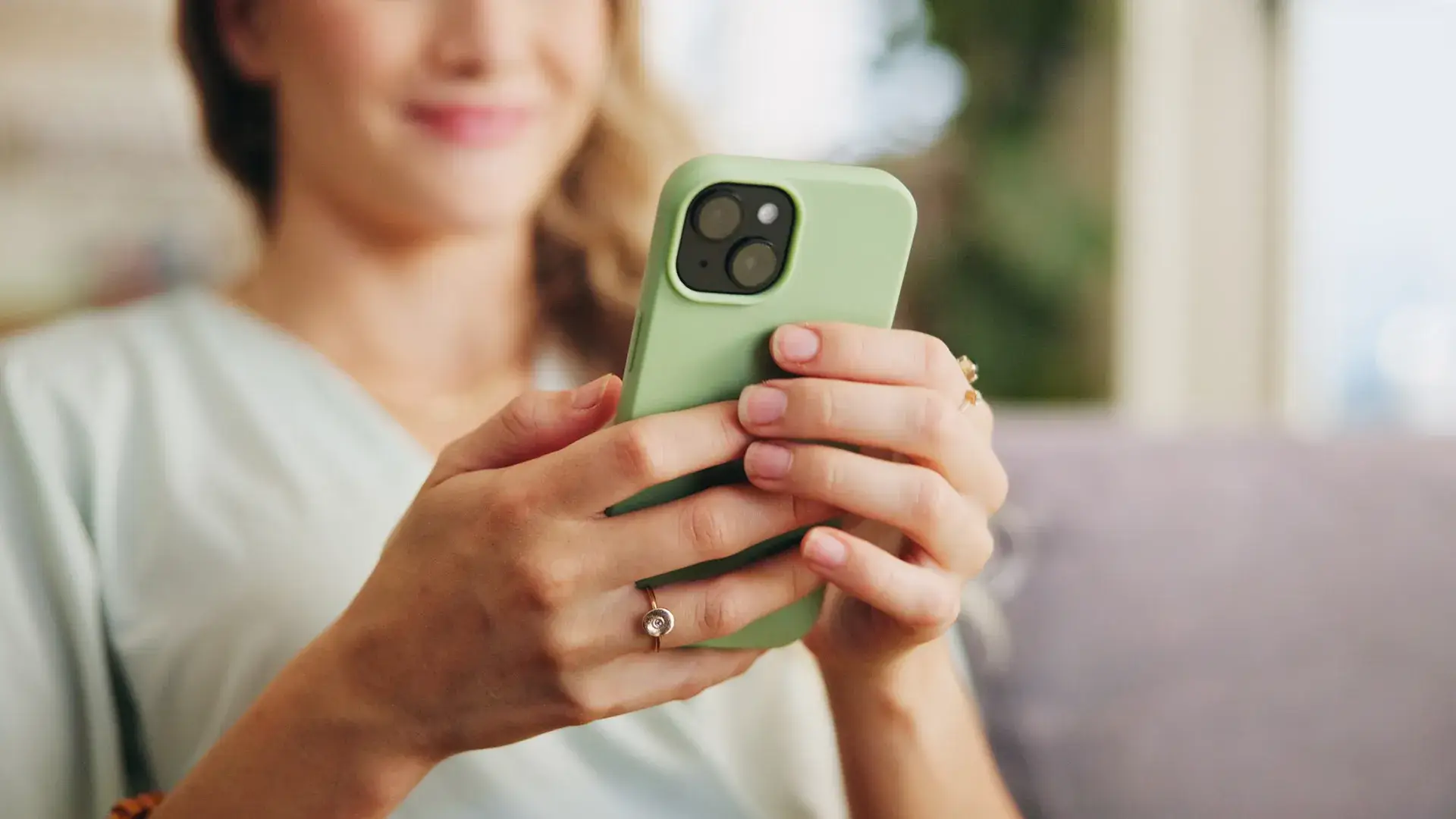 Mujer sosteniendo un smartphone con funda verde sonr&iacute;e mientras lo usa.
