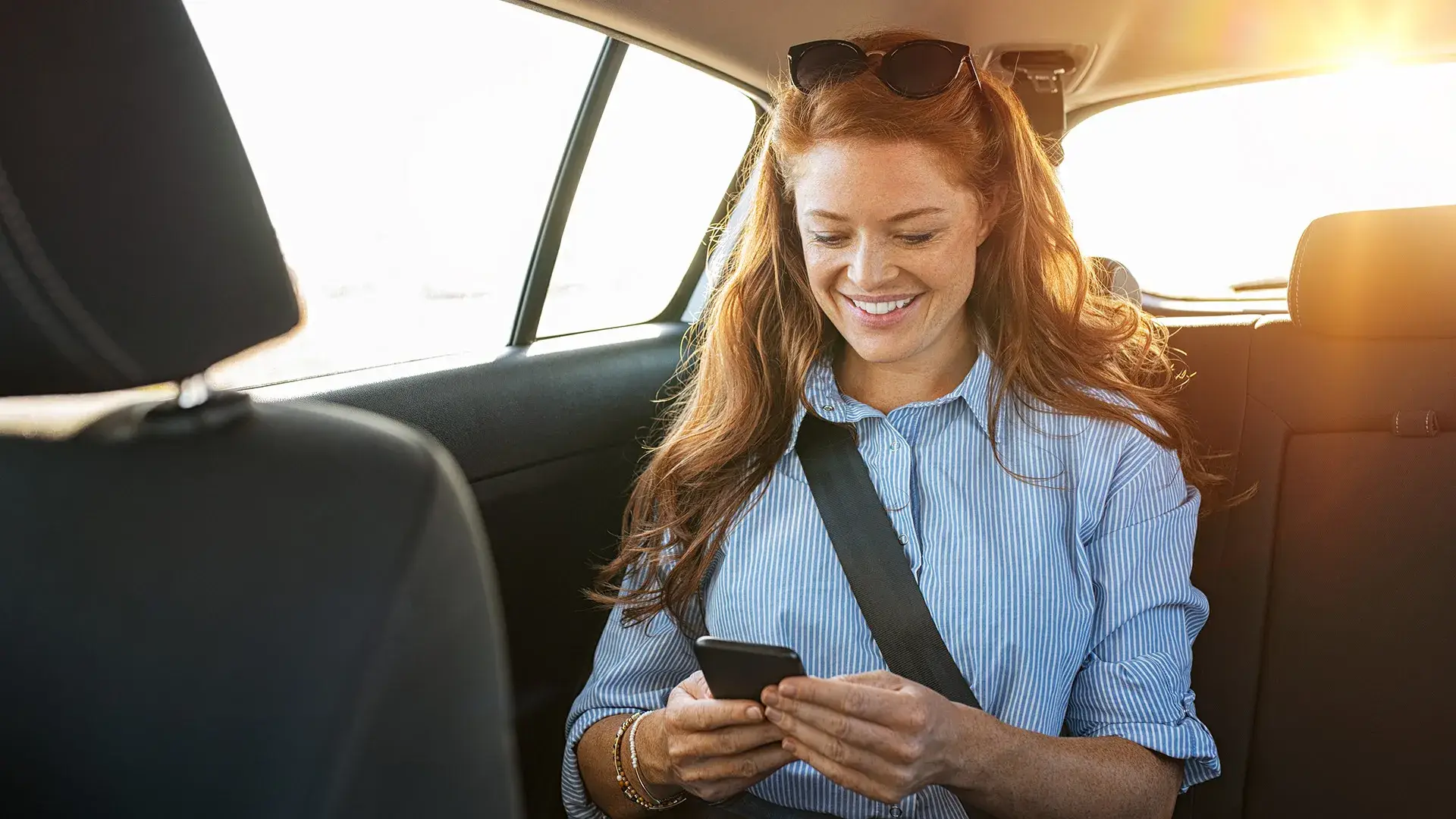 Mujer sonriendo mientras usa su tel&eacute;fono en el asiento trasero de un auto.