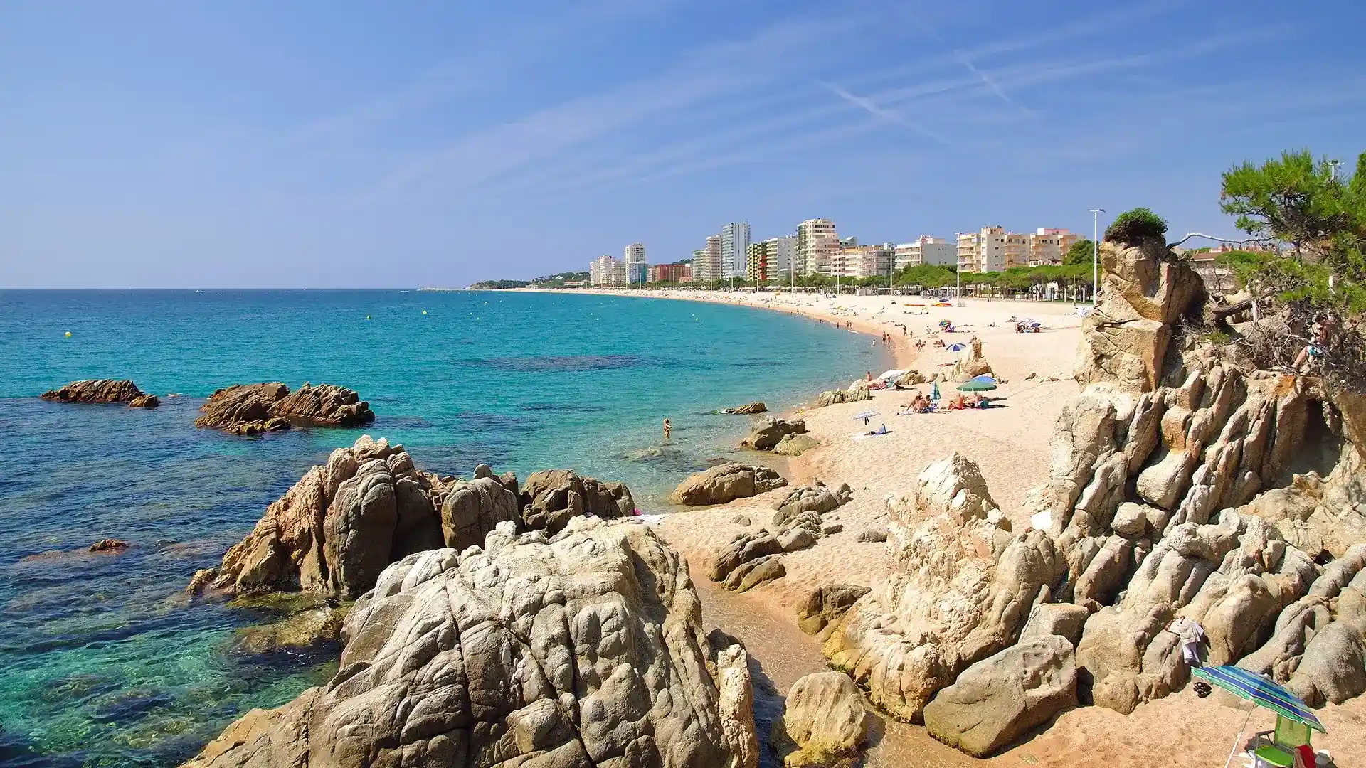 Playa con rocas, mar turquesa y edificios al fondo bajo un cielo azul.