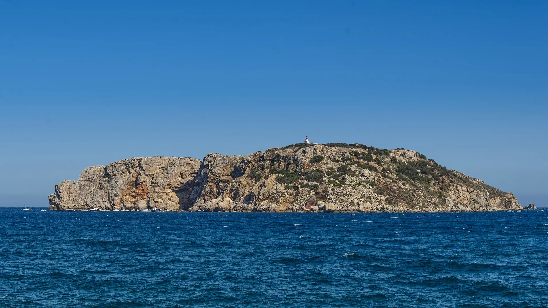 Isla rocosa con faro bajo el cielo azul y mar agitado.