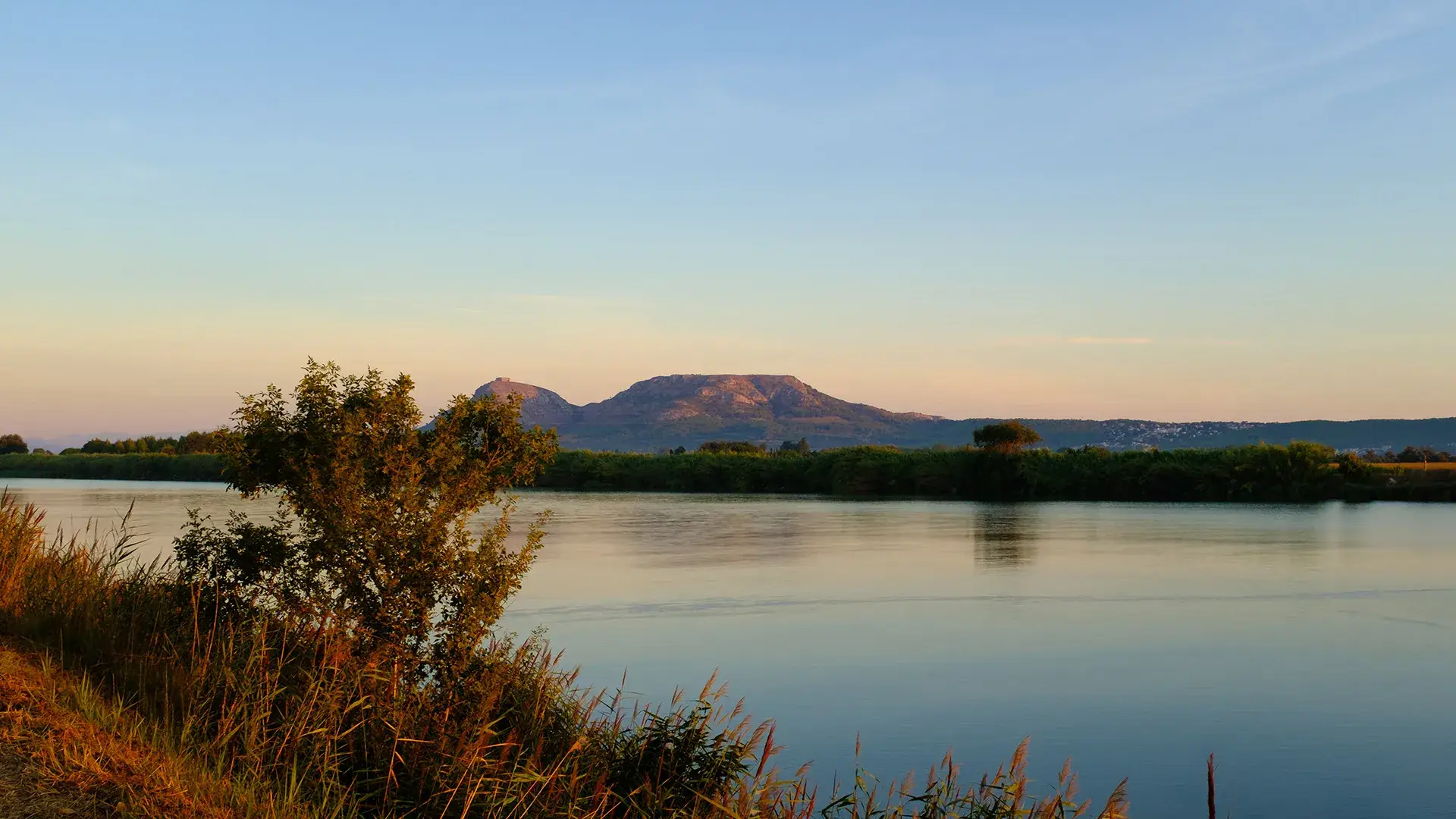 Paisaje al atardecer con lago, monta&ntilde;as y vegetaci&oacute;n al frente.