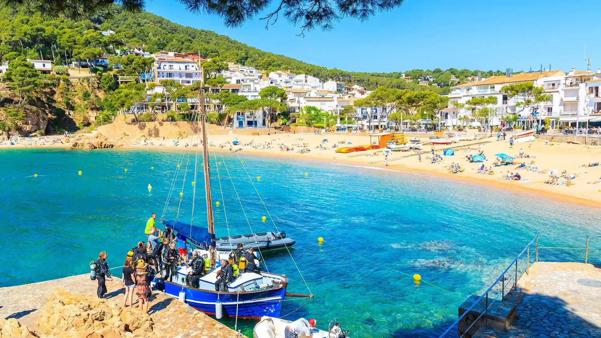 Barco y buceadores en un muelle frente a una playa soleada y casas blancas.