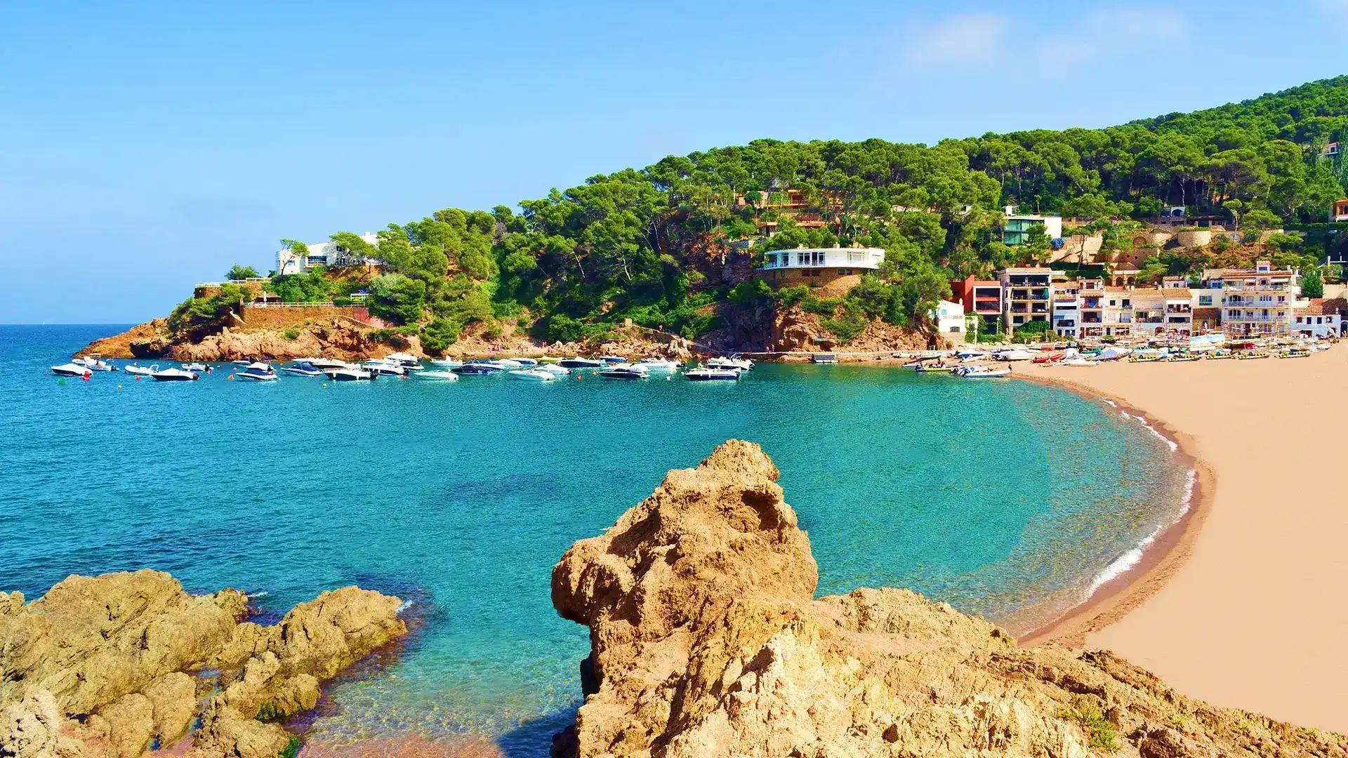 Bah&iacute;a con playa de arena, agua turquesa y casas blancas entre &aacute;rboles verdes.