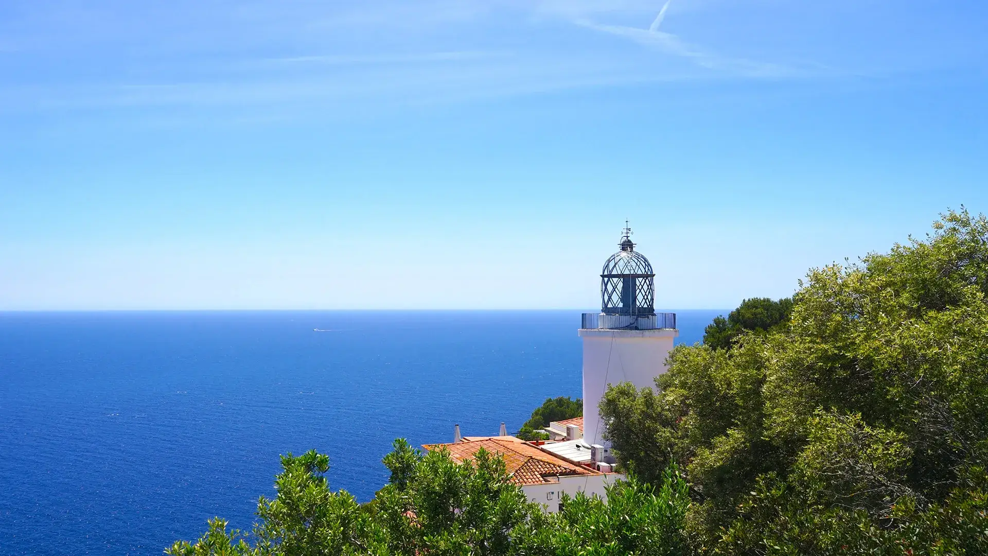 Faro blanco frente al mar azul y cielo despejado, rodeado de vegetaci&oacute;n.