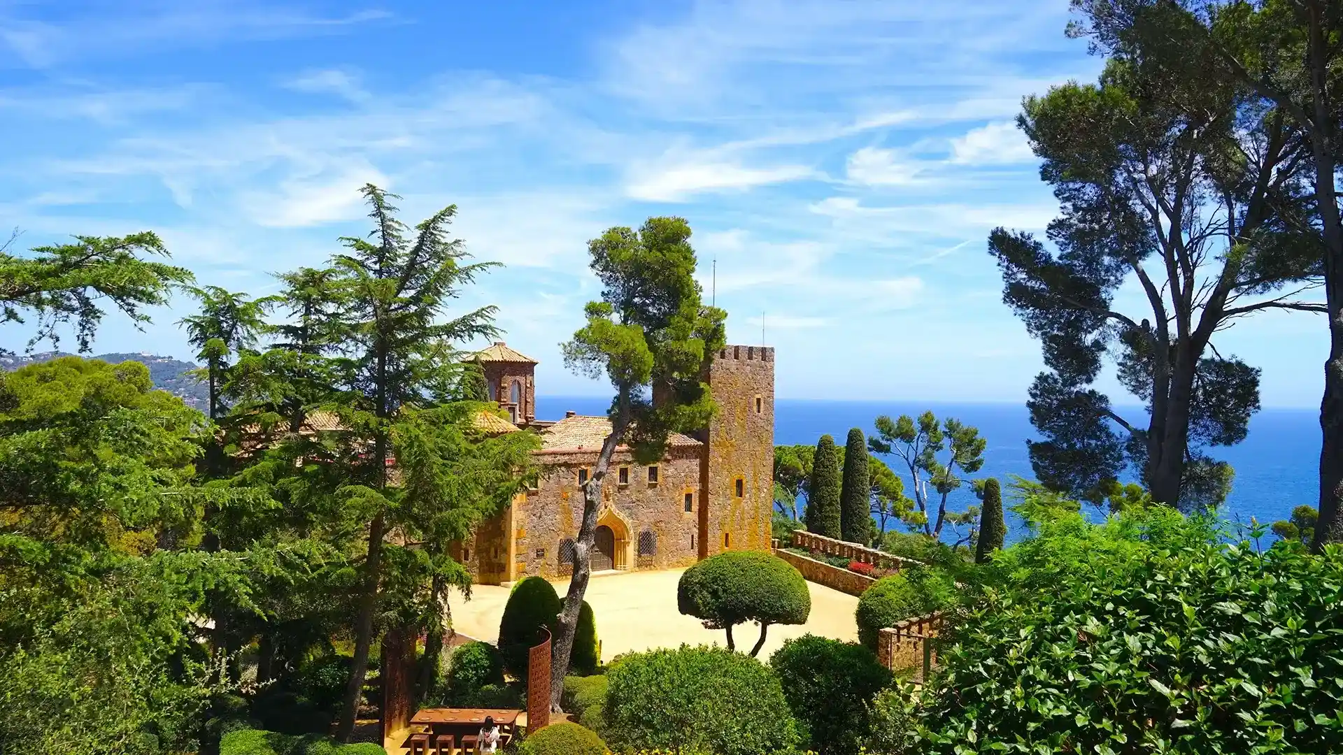 Castillo rodeado de &aacute;rboles con vista al mar azul bajo un cielo despejado.