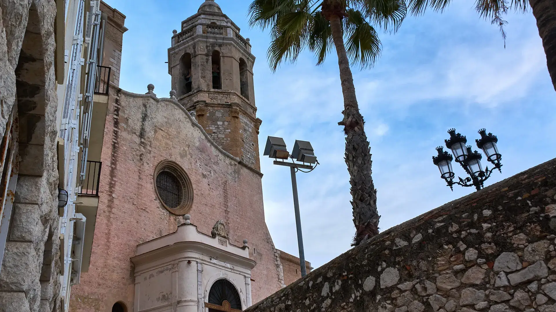 Iglesia de piedra con campanario y palmera bajo un cielo azul.