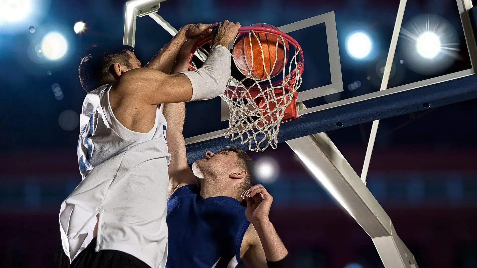 Jugadores de baloncesto disputan la pelota cerca del aro iluminado.
