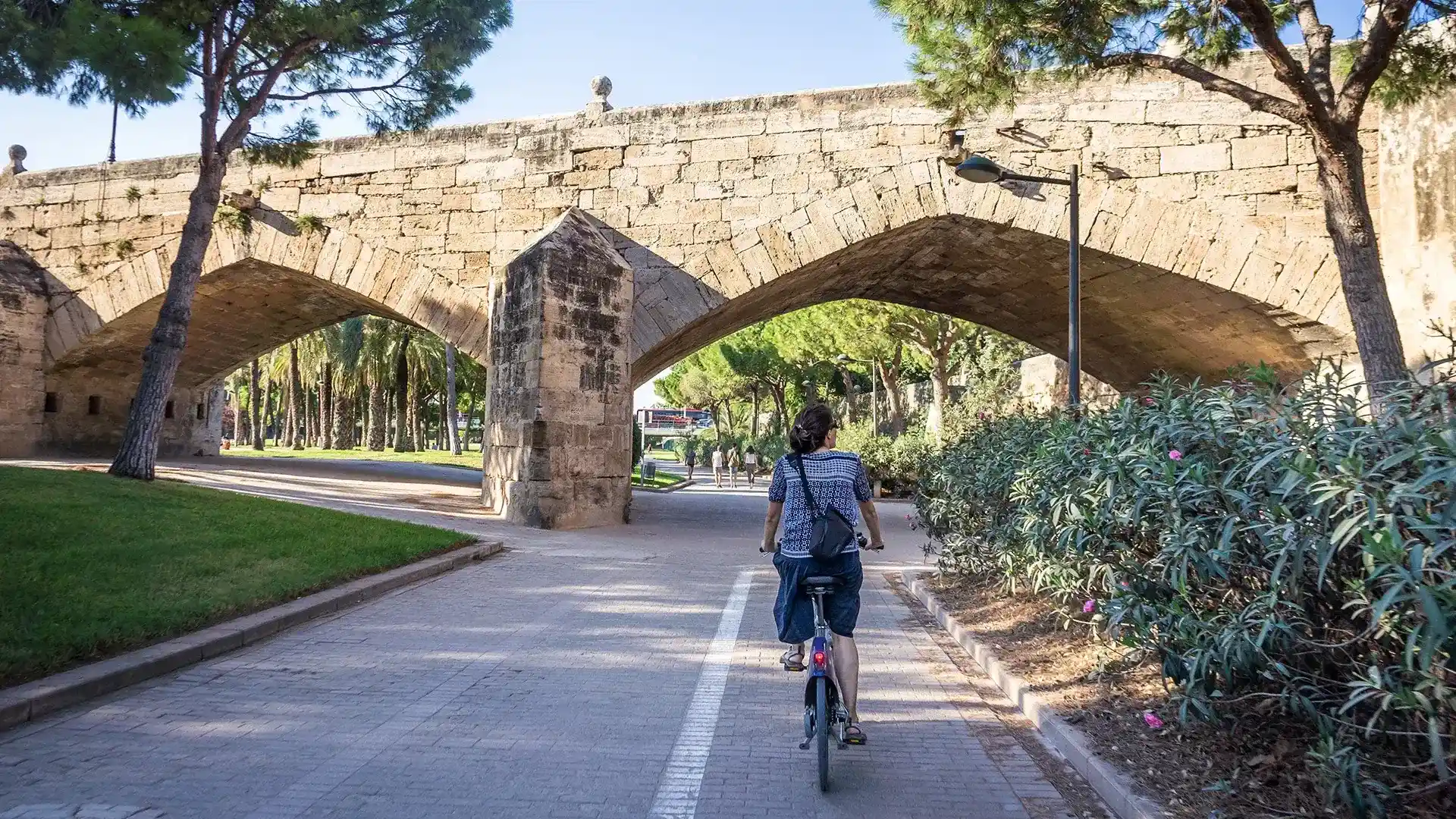 Persona en bicicleta bajo un antiguo puente de piedra rodeado de vegetaci&oacute;n.