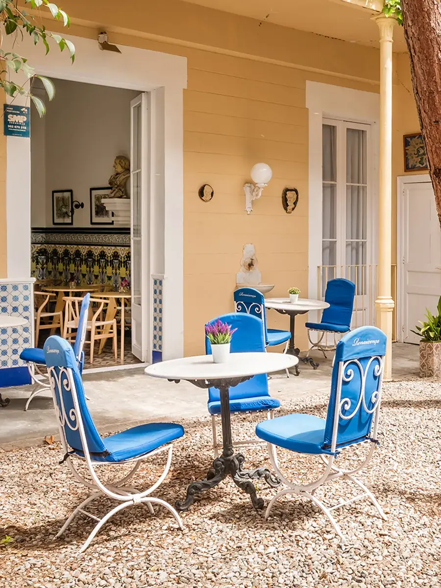 Patio con sillas azules y mesa blanca sobre suelo de piedras, puerta abierta.