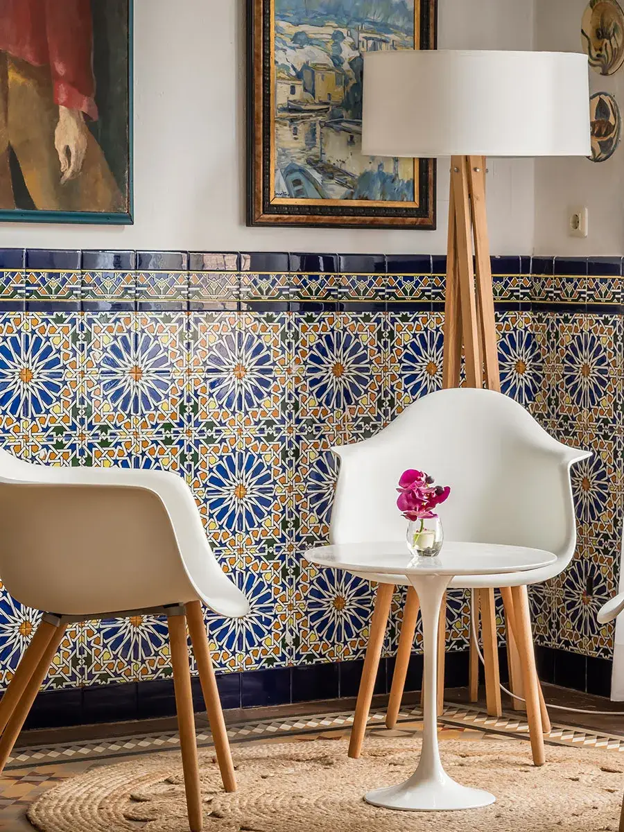 Sala con sillas blancas, mesa peque&ntilde;a, azulejos decorativos y cuadros en la pared.