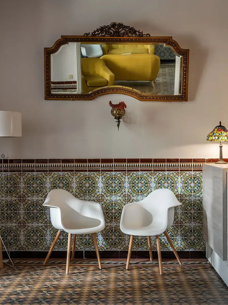 Dos sillas blancas frente a azulejos decorativos y un espejo con marco tallado.