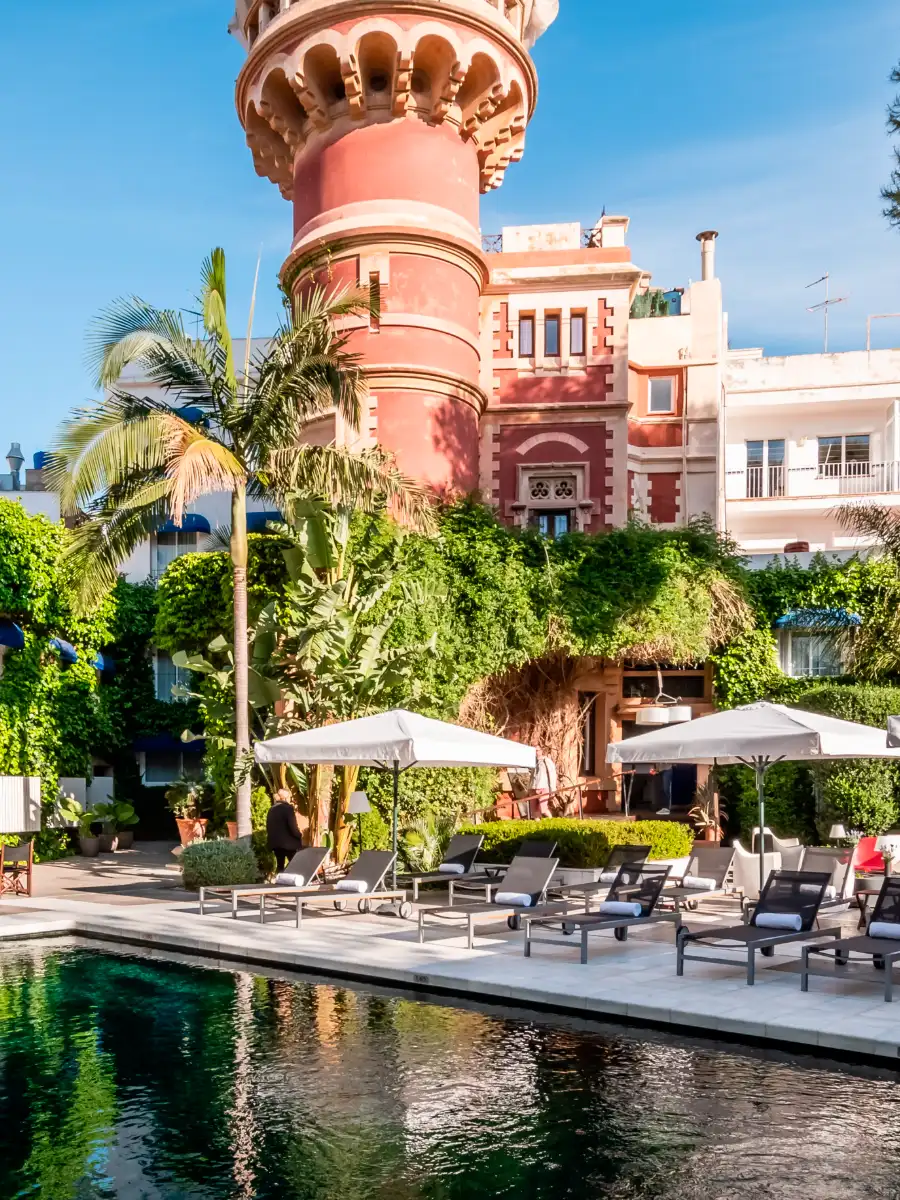 Jard&iacute;n tropical con piscina y edificio de tonos rosados, palmeras y tumbonas
