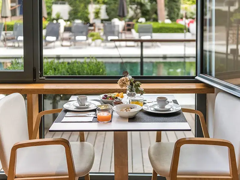 Desayuno servido en mesa junto a gran ventana con vista a la piscina.