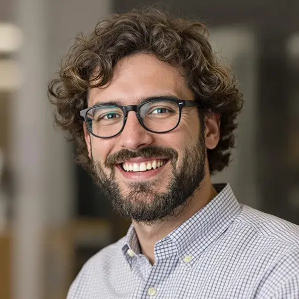 Persona sonriendo, con barba y gafas, fondo interior desenfocado.