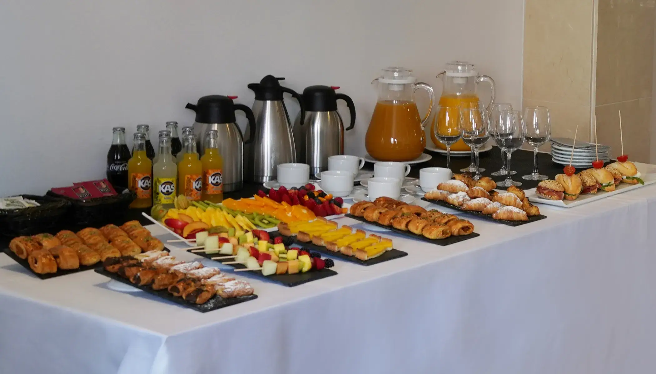 Buf&eacute; con bebidas, frutas, s&aacute;ndwiches y pasteles sobre mantel blanco.