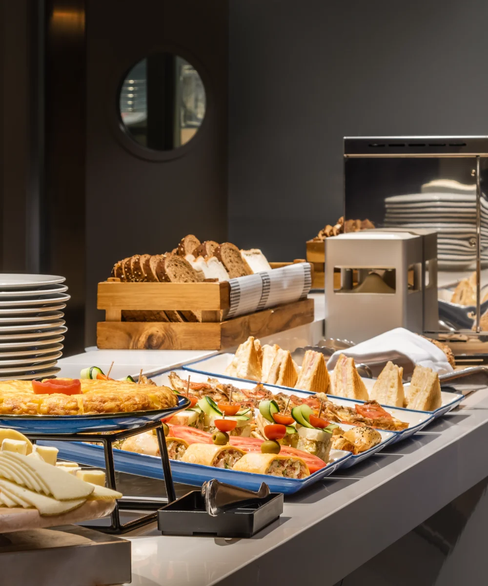 Mesa buffet con panes, quesos y tostadas, bandejas azules y fogones calientes