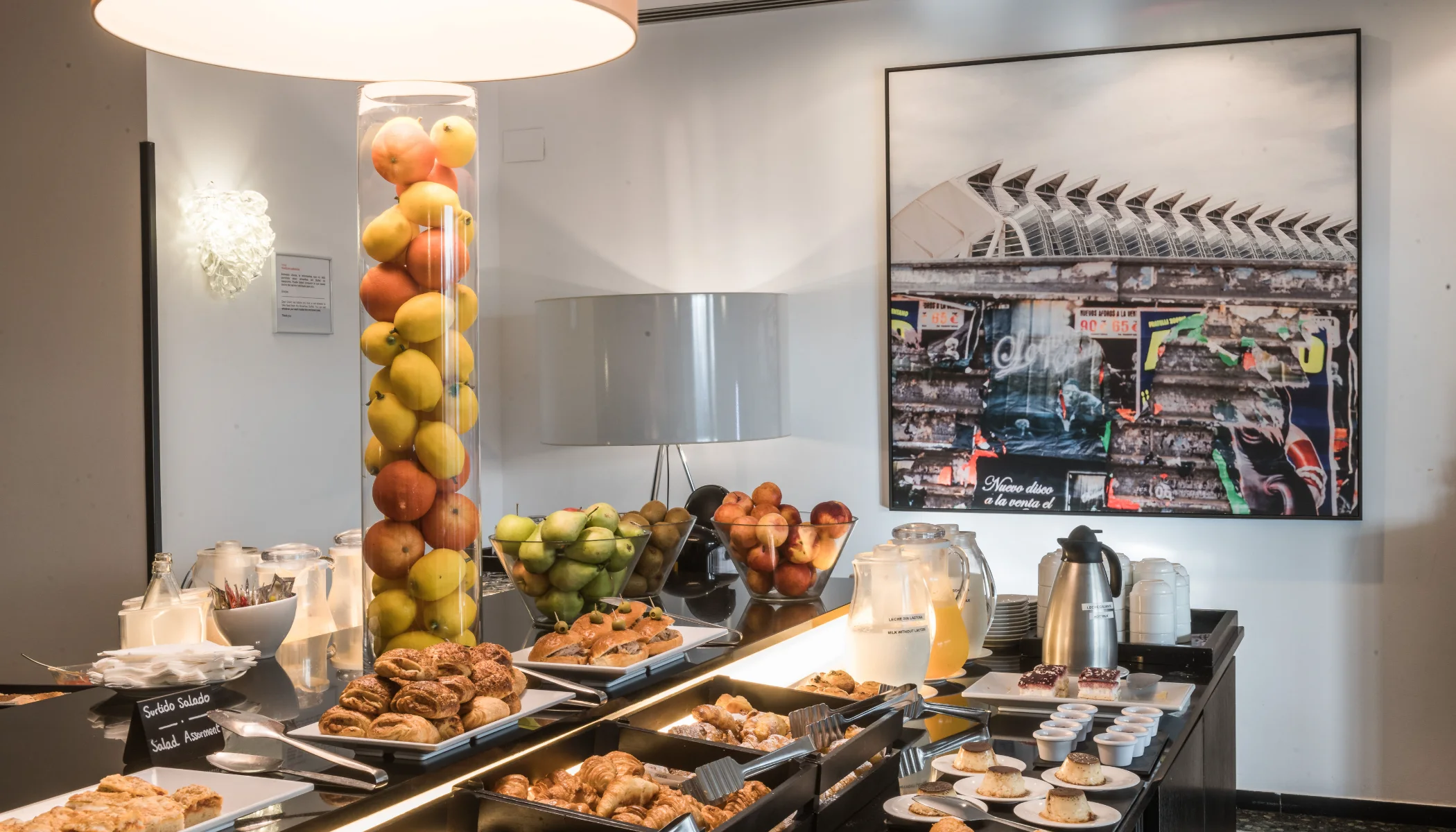 Buffet con frutas, croissants, panecillos y bebidas en un ambiente moderno.
