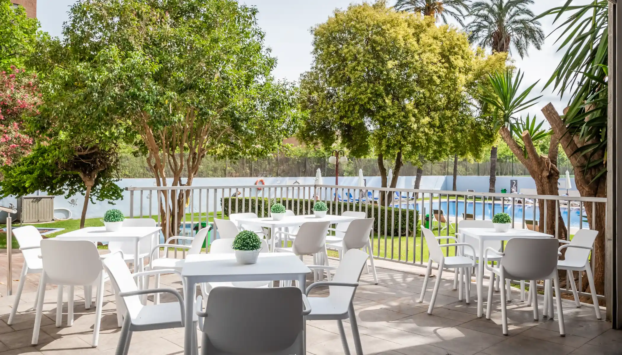 Terraza con mesas blancas y vista a la piscina rodeada de &aacute;rboles.
