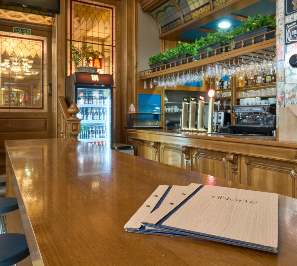 Bar con barra de madera, men&uacute;s y vitrinas con bebidas y copas.