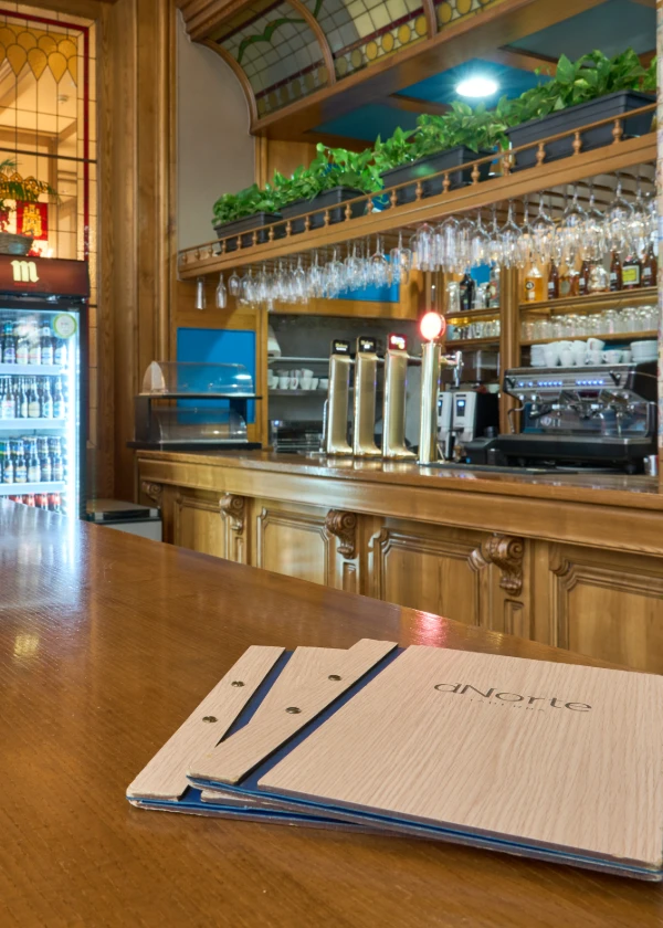 Bar con men&uacute;s sobre la barra y estantes con copas y plantas en el fondo.