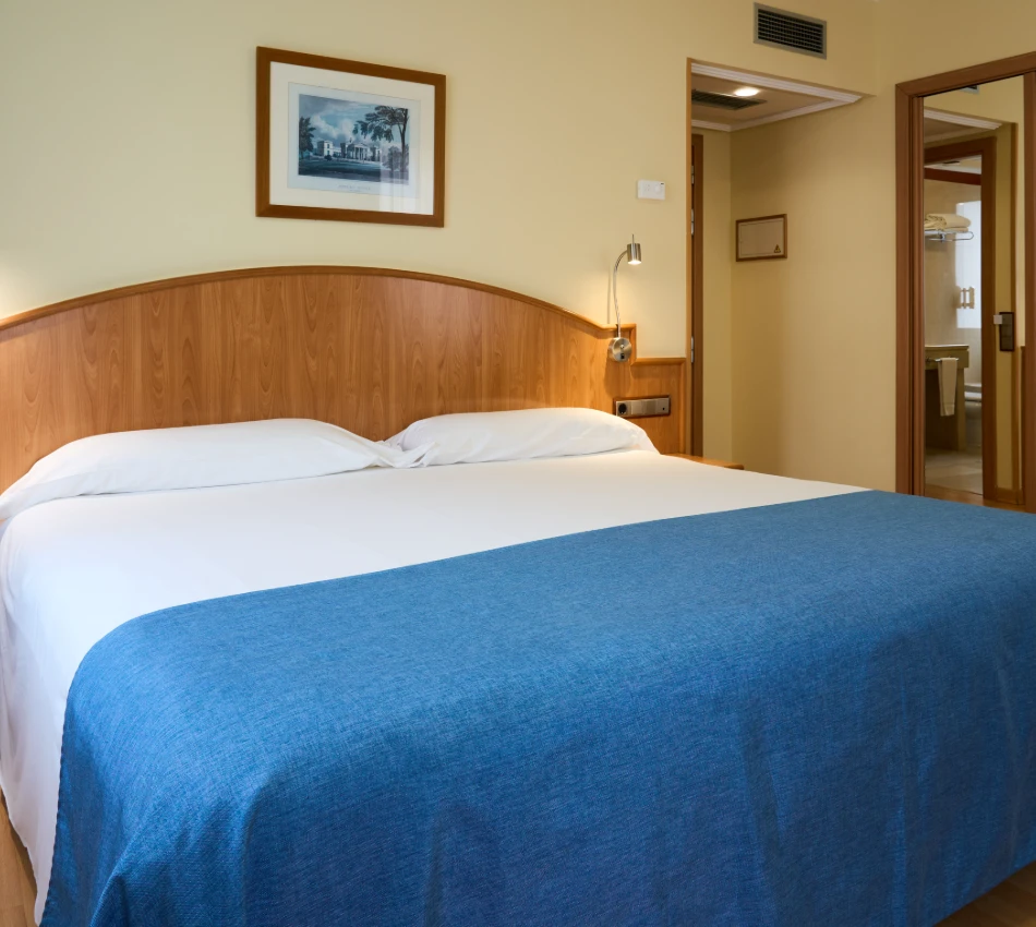 Cama doble en habitaci&oacute;n de hotel con colcha azul y decoraci&oacute;n sencilla.