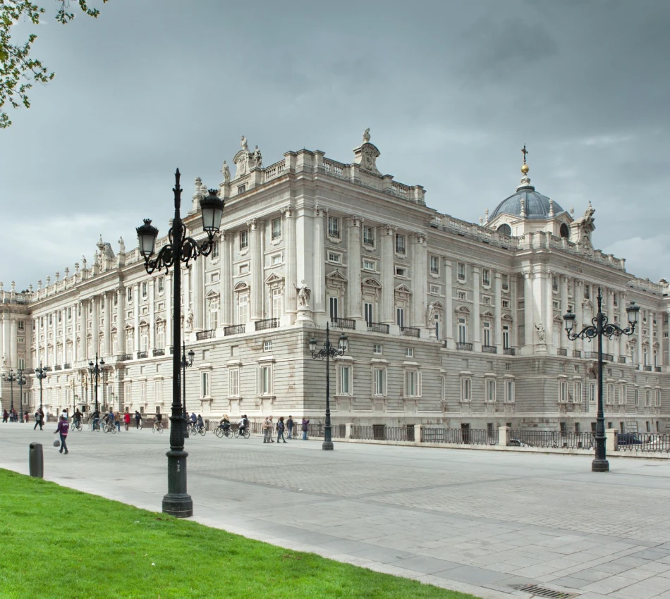 Palacio real con nubes grises, personas caminando y zona verde.