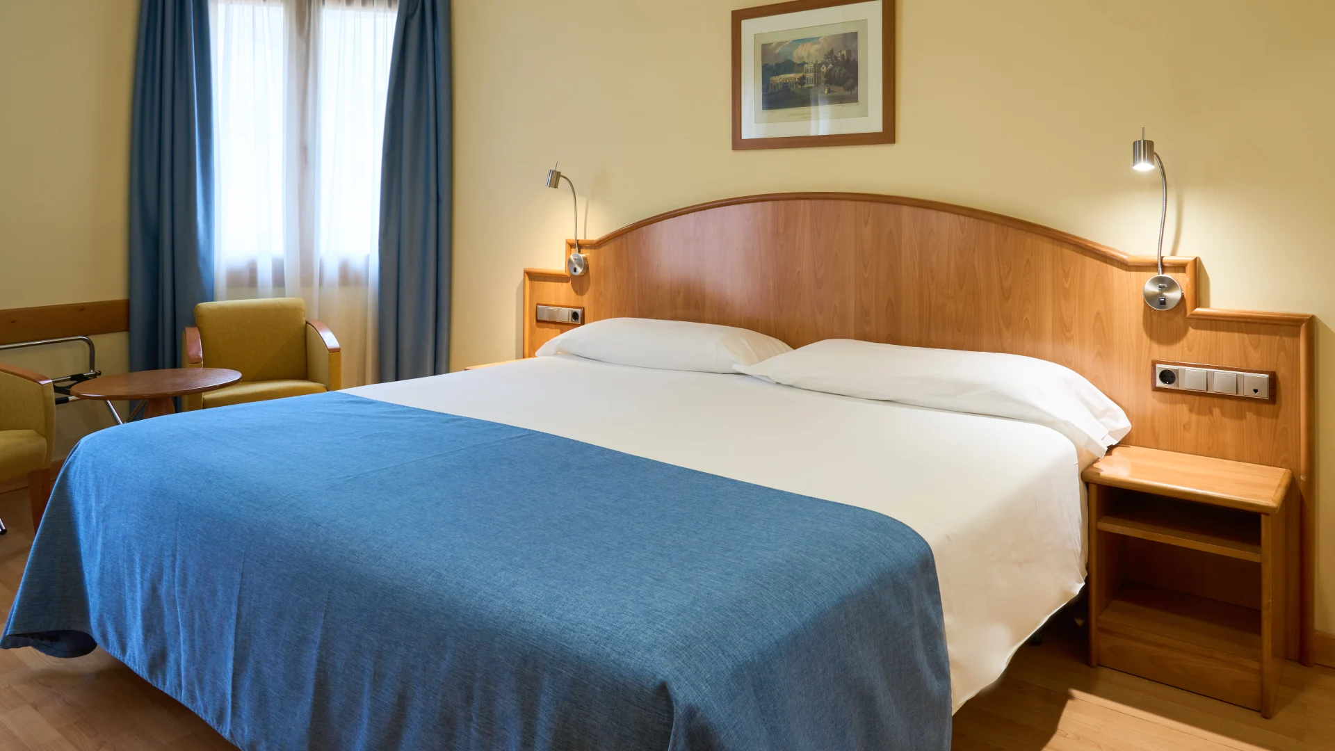 Cama doble con cobertor azul en habitaci&oacute;n de hotel con sillas y mesa peque&ntilde;a.