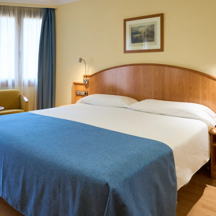 Cama doble con cobertor azul en habitaci&oacute;n iluminada, cuadro y sill&oacute;n.