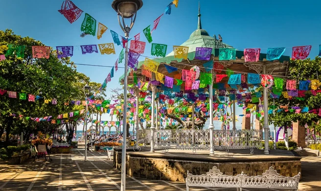 Kiosco decorado con papel picado de colores en una plaza soleada.
