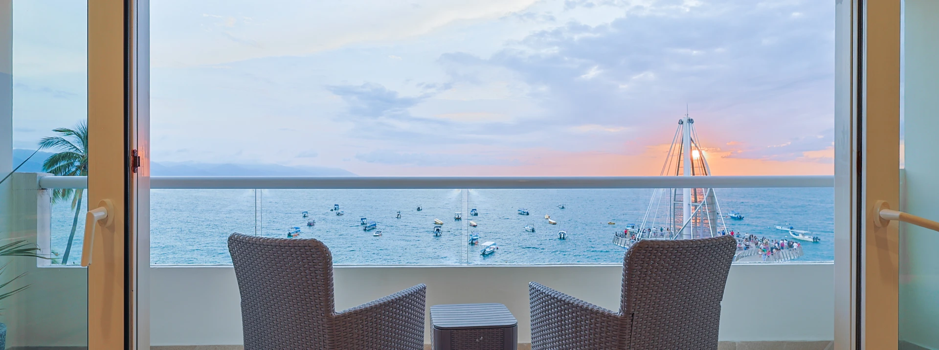 Balc&oacute;n con vista al mar al atardecer, sillas y muelle al fondo.