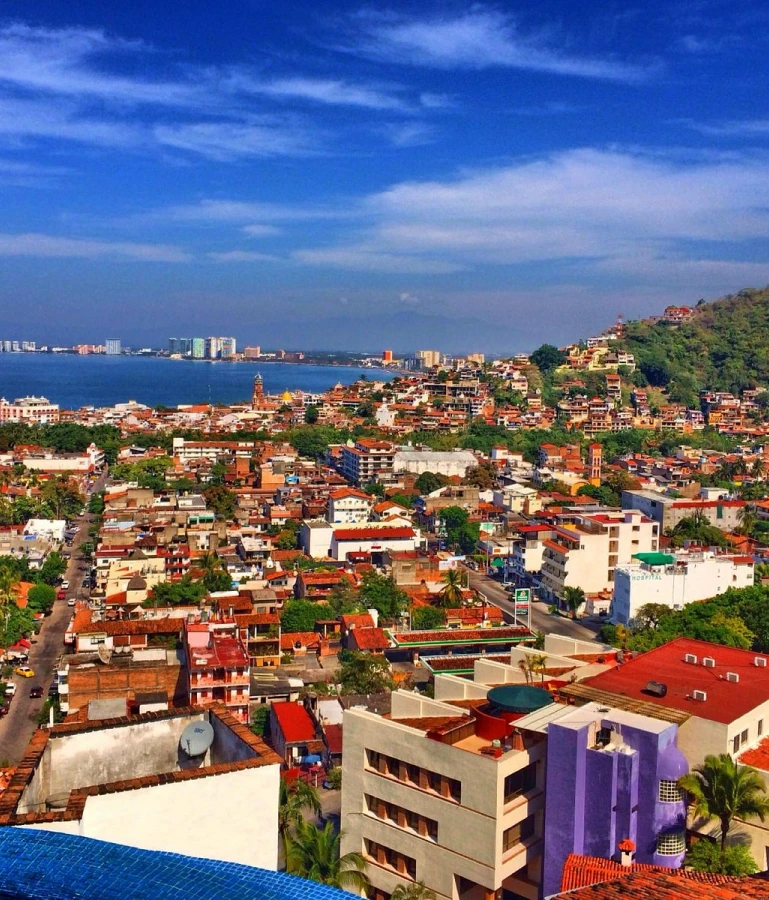 Vista panor&aacute;mica de una ciudad costera con edificios y colinas bajo un cielo azul.