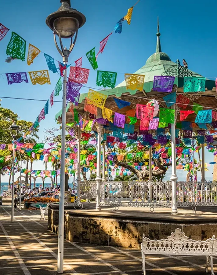 Plaza con quiosco y papel picado multicolor bajo un cielo azul.